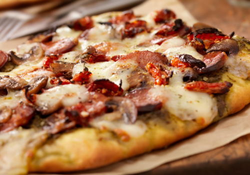 A close-up of a delicious flatbread pizza topped with sausage, mushrooms, cheese, and sun-dried tomatoes on parchment paper.