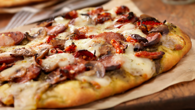 A close-up of a delicious flatbread pizza topped with sausage, mushrooms, cheese, and sun-dried tomatoes on parchment paper.