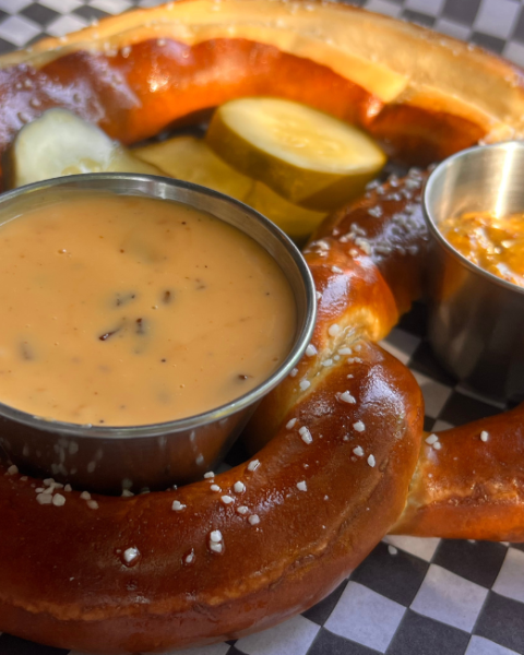 A large pretzel on checkered paper with cheese sauce, mustard, and pickles in small cups, sprinkled with salt.