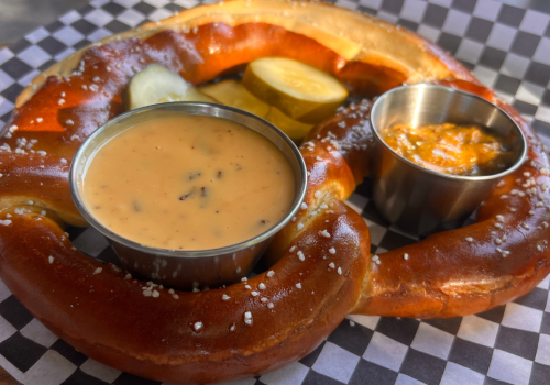 A large pretzel on checkered paper with cheese sauce, mustard, and pickles in small cups, sprinkled with salt.