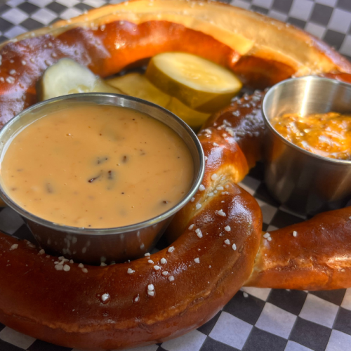A large pretzel on checkered paper with cheese sauce, mustard, and pickles in small cups, sprinkled with salt.