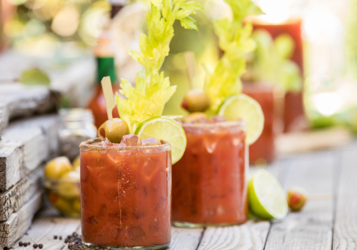 Two Bloody Mary cocktails garnished with celery, olives, and lime slices on a wooden surface.