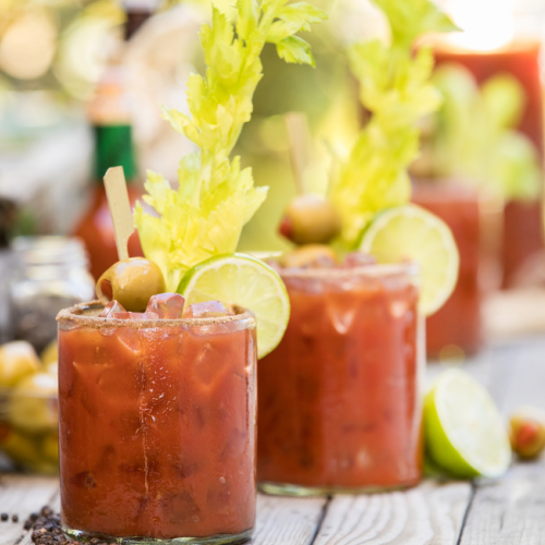 Two Bloody Mary cocktails garnished with celery, olives, and lime slices on a wooden surface.