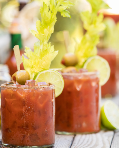Two Bloody Mary cocktails garnished with celery, olives, and lime slices on a wooden surface.