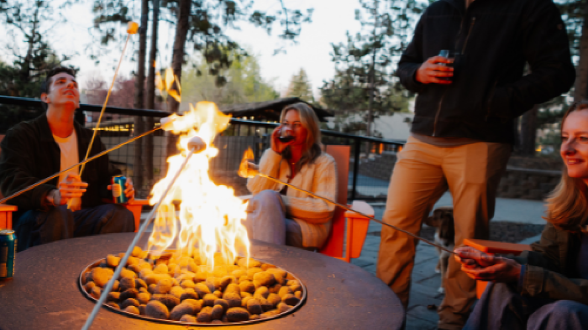 A group of people is gathered around a fire pit, roasting marshmallows and enjoying drinks outdoors at Riverhouse Lodge in Bend Oregon