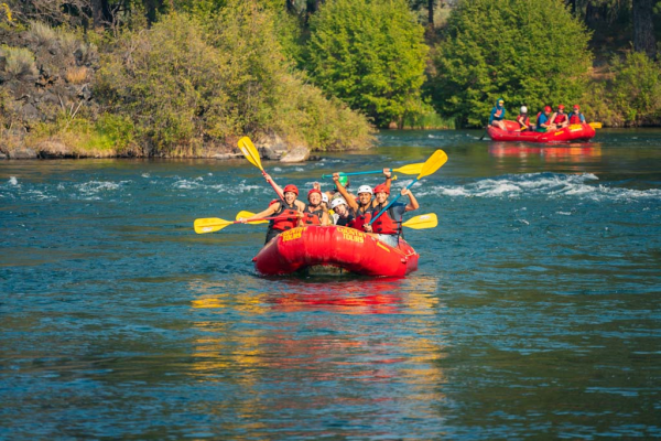 People rafting in red boats with yellow paddles on a river, surrounded by green trees and blue water, enjoying outdoor adventure fun.