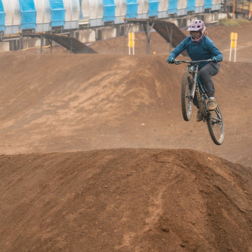 A person is mountain biking, performing a jump on a dirt track, wearing a helmet and protective gear.