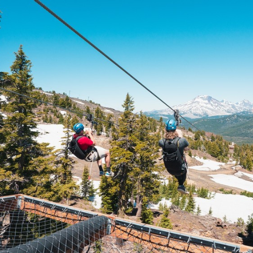 Two people are ziplining between trees on a mountainside with snow patches and a distant mountain view under a clear blue sky.