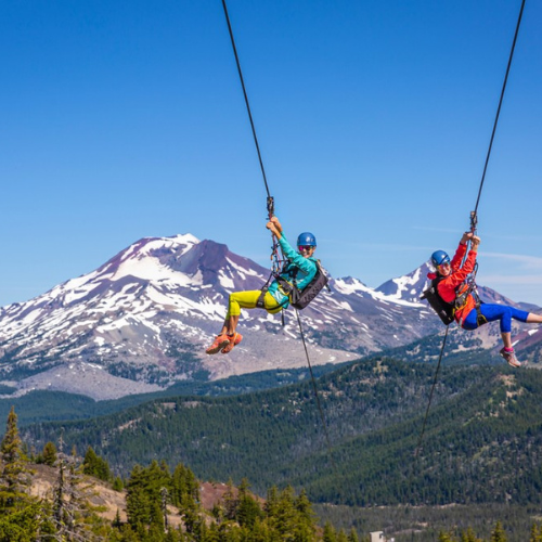 Two people zip-lining with a scenic mountain view in the background and clear blue skies.