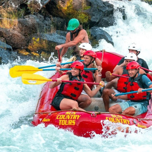 A group of people wearing helmets and life jackets are whitewater rafting in a red raft down a rocky river.
