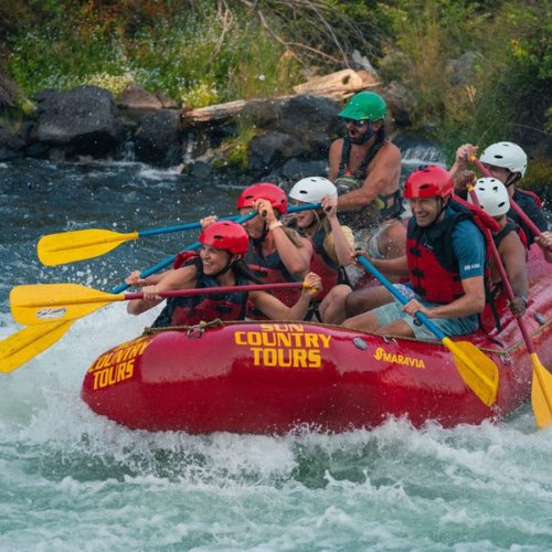 A group of people are white-water rafting in a red raft with yellow paddles, wearing helmets and life vests, navigating through rapids.