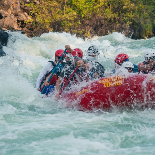 A group of people is whitewater rafting on a turbulent river, wearing helmets and life vests, on a red inflatable raft.