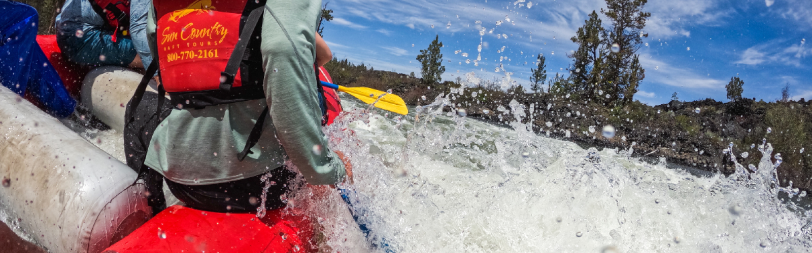 People are whitewater rafting on a river, wearing life jackets, with water splashing around them against a backdrop of trees and sky.