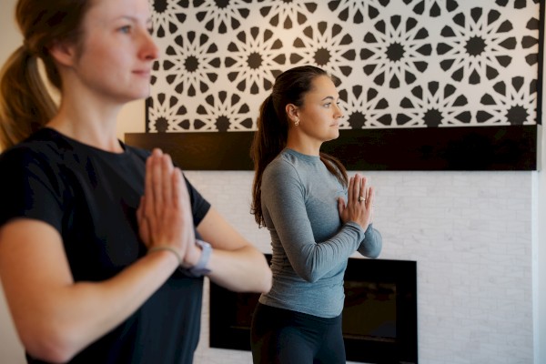 Two individuals are practicing yoga at Riverhouse Lodge in Bend, Oregon, standing in a meditative pose with their hands in prayer position, facing forward.