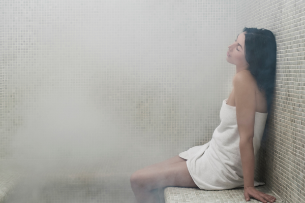 A person wrapped in a towel relaxes in a steamy sauna at Riverhouse Lodge in Bend, Oregon, sitting against a tiled wall with eyes closed.