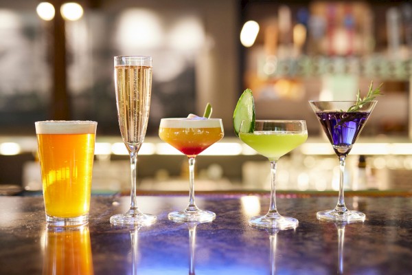 An array of drinks on a bar counter at Currents in Bend, Oregon: a beer, champagne, and colorful cocktails garnished with lime and herbs, in a dimly lit setting.