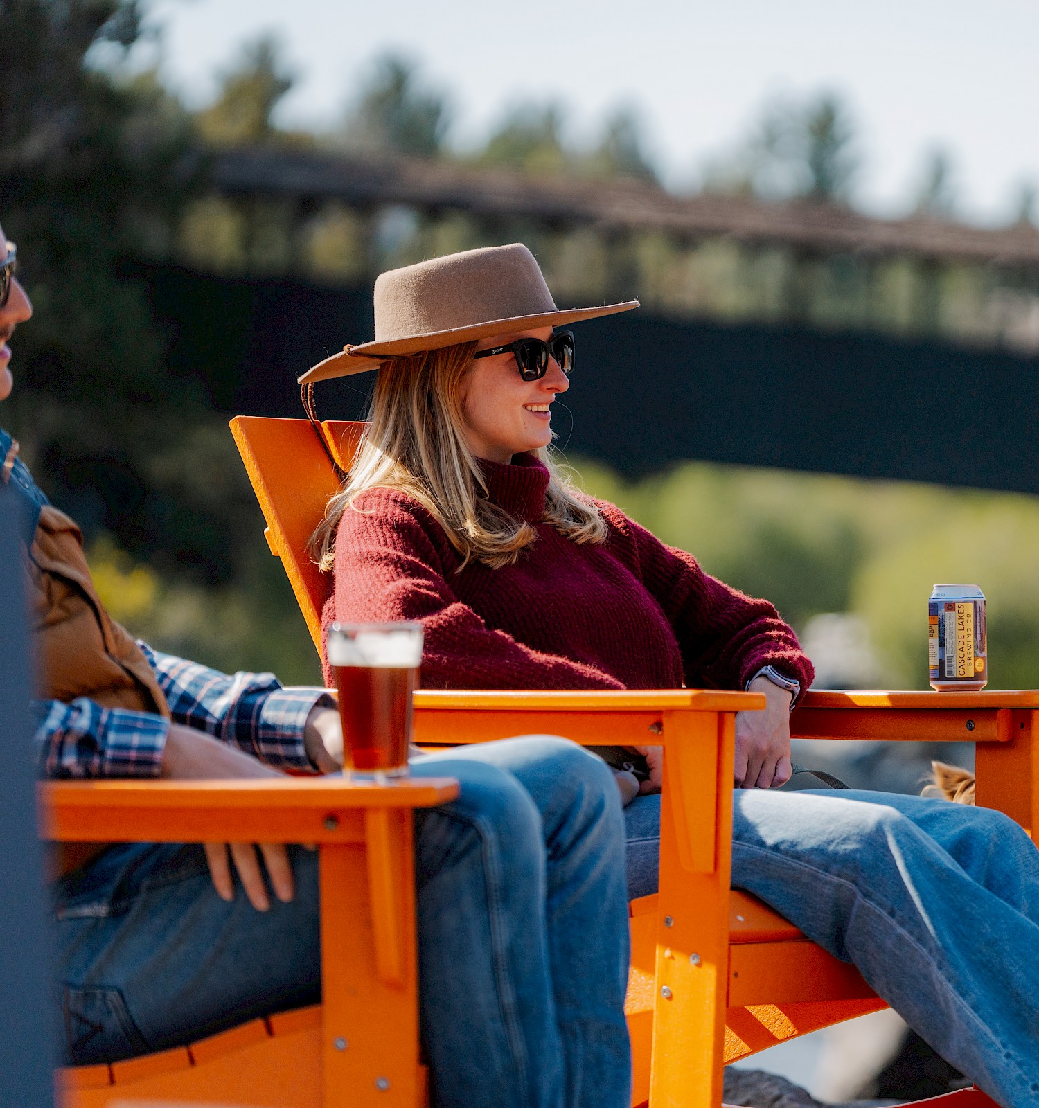 People are relaxing outdoors in orange chairs, enjoying drinks with a scenic bridge and trees in the background. Stay at Riverhouse Lodge in Bend, Oregon for the best located hotel along river in Bend, Oregon