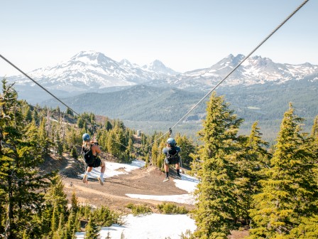 Two people zip-lining over a scenic forest landscape with snow-capped mountains in the background under a clear blue sky.