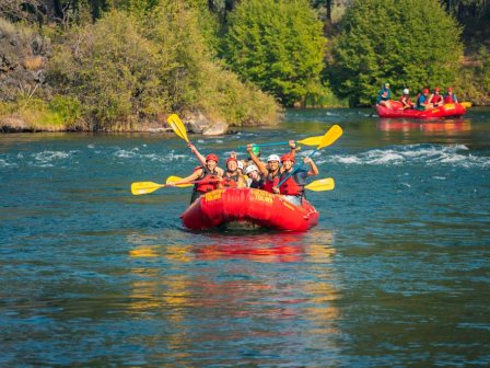People rafting in red boats with yellow paddles on a river, surrounded by green trees and blue water, enjoying outdoor adventure fun.