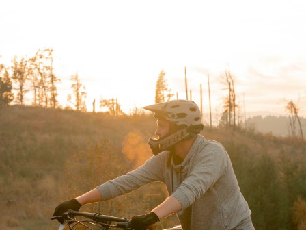 A person wearing a helmet and gloves is standing with their mountain bike in a scenic outdoor area during sunset.