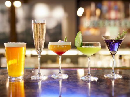 An array of drinks on a bar counter at Currents in Bend, Oregon: a beer, champagne, and colorful cocktails garnished with lime and herbs, in a dimly lit setting.