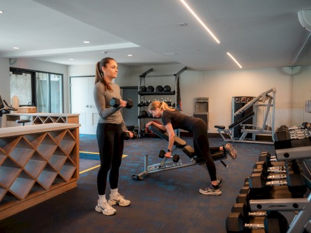 Two people are exercising in the gym at Riverhouse Lodge in Bend Oregon, using dumbbells and fitness equipment. The room is well-lit with various workout machines.