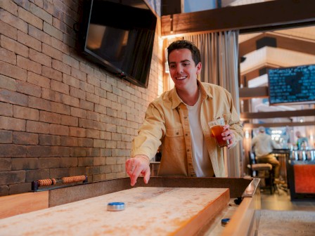 A person is playing shuffleboard at Currents at Riverhouse Lodge in Bend, Oregon, holding a drink. They are in a casual setting with a TV and menu board in the background.