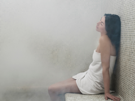 A person wrapped in a towel relaxes in a steamy sauna at Riverhouse Lodge in Bend, Oregon, sitting against a tiled wall with eyes closed.