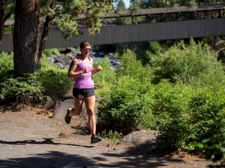 A person in athletic attire runs on a shaded trail with greenery and a bridge visible in the background, enjoying an outdoor workout.