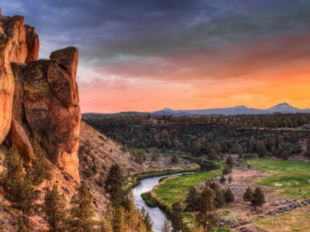 The image showcases a scenic view with a large rock formation, a winding river, fields, and a colorful sunset sky, appearing serene and picturesque.