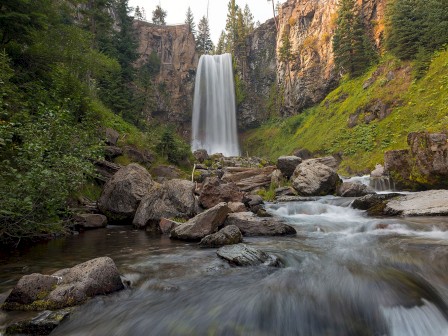 A cascading waterfall descends into a rocky stream, surrounded by lush greenery and tall cliffs, creating a serene natural scene.