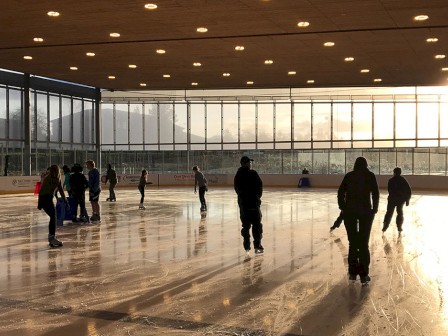 People are ice skating inside a bright, sunlit ice rink with large windows and a wooden ceiling.