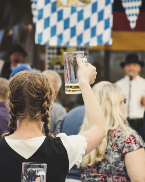 People at a festival, some holding beer mugs, with German-themed decorations like flags and attire visible in the background.