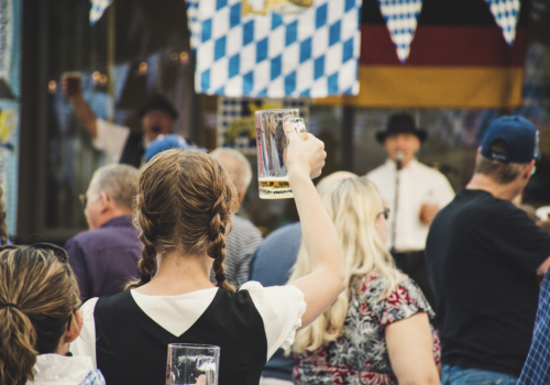 People at a festival, some holding beer mugs, with German-themed decorations like flags and attire visible in the background.