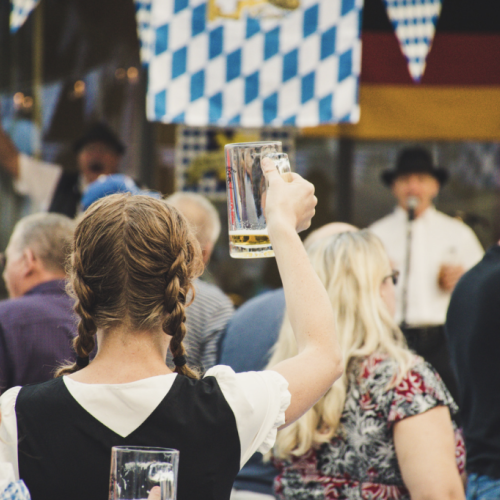 People at a festival, some holding beer mugs, with German-themed decorations like flags and attire visible in the background.