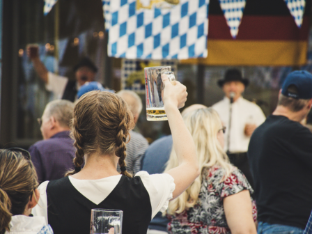 A crowd enjoying a festive event; a woman raises a beer mug, with Bavarian flags and a musician visible in the background.