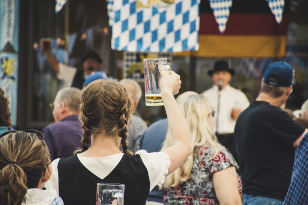 A crowd enjoying a festive event; a woman raises a beer mug, with Bavarian flags and a musician visible in the background.