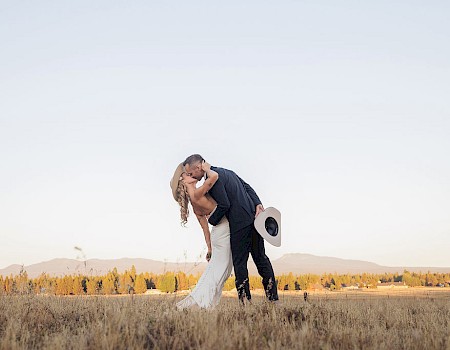A couple is kissing in a field during sunset, with the man holding a hat, and mountains visible in the background.