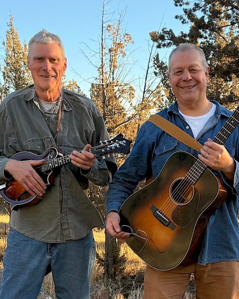 Live Americana, folk, and bluegrass music in Bend Oregon – The Mavis Bretz Project playing guitar and mandolin at Currents at the Riverhouse, September 5, 2025. Two musicians smiling outdoors with trees and sunny sky in the background