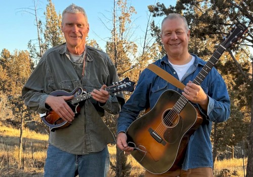 Live Americana, folk, and bluegrass music in Bend Oregon – The Mavis Bretz Project playing guitar and mandolin at Currents at the Riverhouse, September 5, 2025. Two musicians smiling outdoors with trees and sunny sky in the background