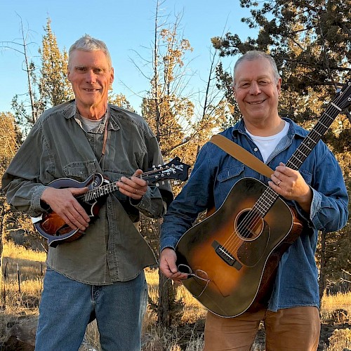 Live Americana, folk, and bluegrass music in Bend Oregon – The Mavis Bretz Project playing guitar and mandolin at Currents at the Riverhouse, September 5, 2025. Two musicians smiling outdoors with trees and sunny sky in the background