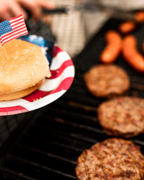 A grill with burgers and sausages, a plate with a bun and a small flag, suggesting a festive barbecue gathering.