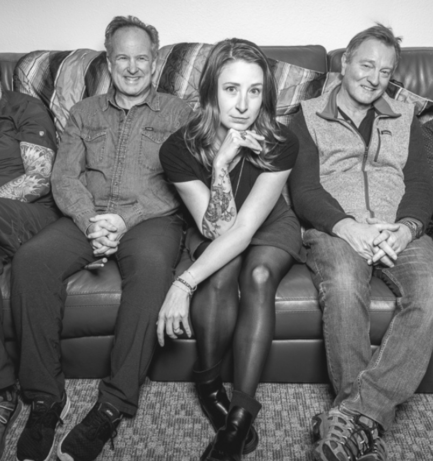 A group of five people sitting on a couch, two holding guitars, the central figure is a woman in front.