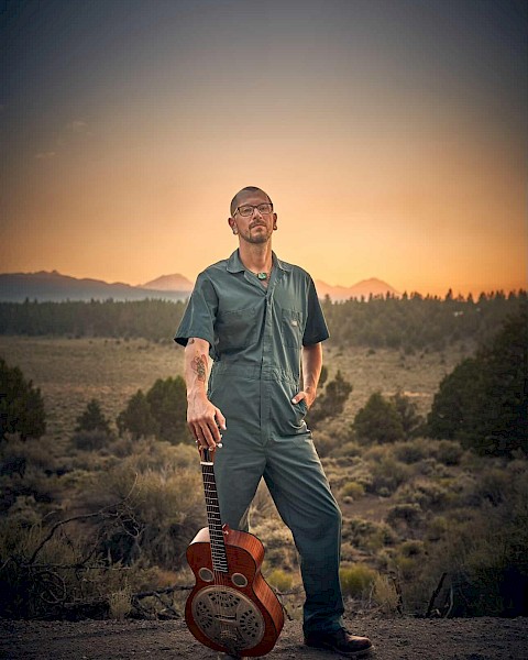 Aaron Elston in a jumpsuit standing in a desert landscape at sunset with a guitar, blues musician performing live at Currents at Riverhouse Lodge in Bend Oregon on October 17, 2025.