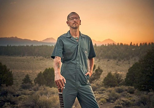 Aaron Elston in a jumpsuit standing in a desert landscape at sunset with a guitar, blues musician performing live at Currents at Riverhouse Lodge in Bend Oregon on October 17, 2025.