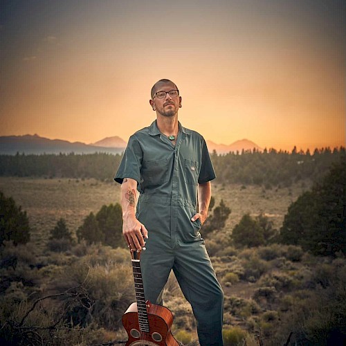 Aaron Elston in a jumpsuit standing in a desert landscape at sunset with a guitar, blues musician performing live at Currents at Riverhouse Lodge in Bend Oregon on October 17, 2025.
