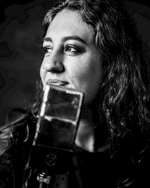 Black and white photo of Brenna Larsen with long hair holding a vintage microphone, thoughtful expression, performing live at Currents at Riverhouse Lodge in Bend Oregon on October 24, 2025