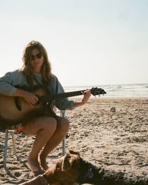 Kara Grace sitting on a beach chair by the ocean playing guitar with a truck nearby, folk singer performing live at Currents at Riverhouse Lodge in Bend Oregon on October 5, 2025