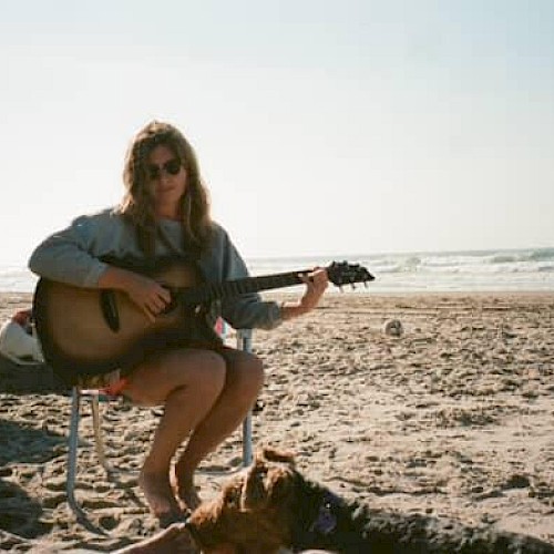Kara Grace sitting on a beach chair by the ocean playing guitar with a truck nearby, folk singer performing live at Currents at Riverhouse Lodge in Bend Oregon on October 5, 2025