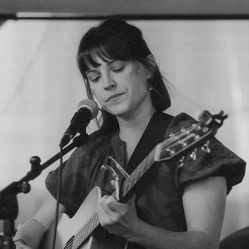 A person is playing an acoustic guitar and singing into a microphone in a black and white photo.